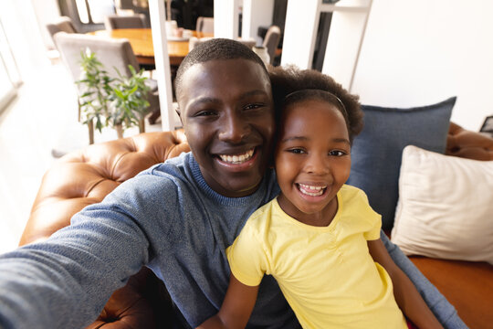 Image of happy african american father and daughter sitting on sofa and taking selfie