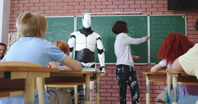 Robot with teen small schoolboy standing in front of class with pupils and teaching robotics. Ed-tech concept. Learning tech at school. Technology for studying. Android teacher and boy.