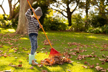 Fototapeta premium Image of happy caucasian boy swiping leaves in autumn garden