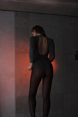 young teen girl in a shiny silver bodysuit is standing fashion from the back with hands near waist and looking down on gray wall background in loft interior with red light. fashion concept, free space