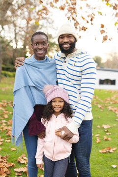 Vertical image of happy african american parents and daughter posing at camera outdoors in autumn