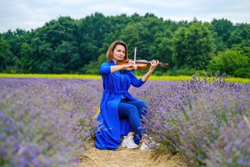 Full body adult woman violinist playing violin and sitting on summer lavender field, romantic...