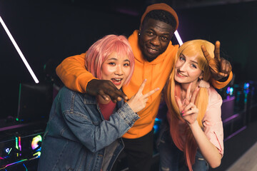 Multiracial young cheerful friends standing close together posing at camera in a dark gaming room with neon lights showing peace and thumbs up signs. High quality photo © PoppyPix