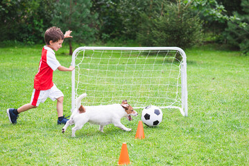 Kid boy in red and white kit playing football (soccer) with his dog at training ground