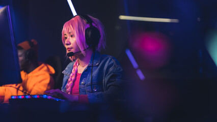 Focused asian female gamer with pink long braid wearing a denim jacket looking at a screen, looking concentrated during an online streaming of a video game. . High quality photo © PoppyPix