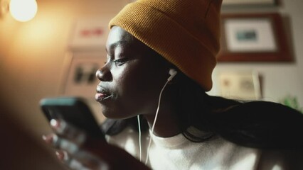 Cheerful African young woman wearing white sweater writing and listening music in headphones in cafe
