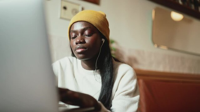 Smiling African young woman wearing white sweater working on laptop and listening music in headphones in cafe