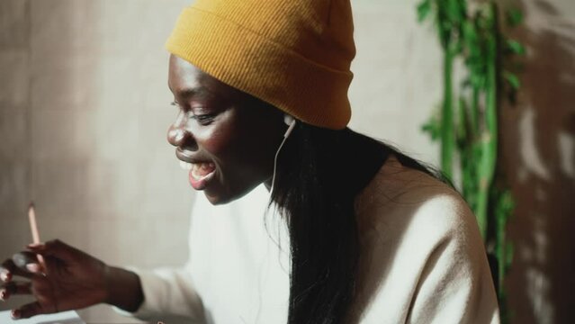 Funny African young woman wearing white sweater talking by video call on laptop in headphones in cafe