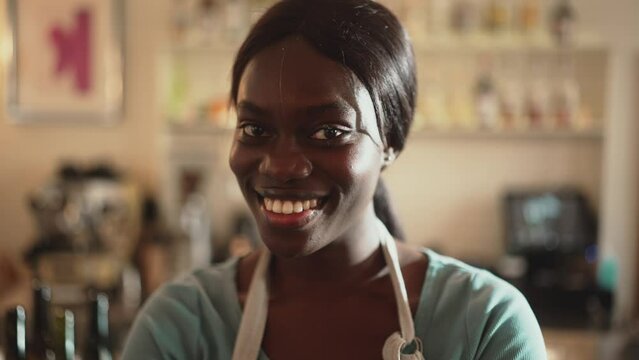 Cheerful African woman barista wearing white apron looking at camera in cafe