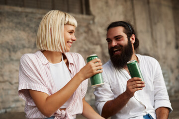 Closeup of young smiling couple toasting with beer cans in an urban environment - 517709667