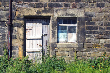 derelict abandoned old stone house with broken window and shabby wooden door overgrown with weeds