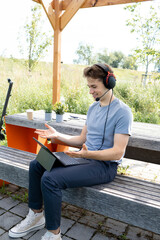 cool, young man with grey shirt sitting outdoor in a park with headset and working on his tablet, in the background e-scooter is standing
