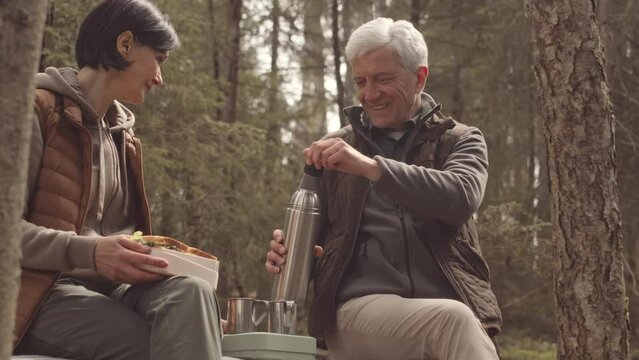 Slowmo of romantic mature couple talking while having lunch in peaceful forest. Man pouring hot tea in thermos mugs