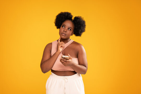 Young Afro Woman Holding Mobile Phone And Thinking About Buying Something Online. Sale Concept. Studio Shot.