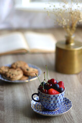 Plate of chocolate chip cookies, cup filled with strawberries, blueberries and cherries, open book and vase with gypsophila flowers on the table. Selective focus.
