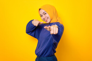 Cheerful young Asian Muslim woman dressed in casual sweater pointing fingers to camera with happy expression isolated over yellow background