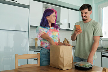 Hipster couple unpacking a paper bag full of take out food containers. Young woman with bright colorful hair and bearded man at the kitchen with delivered food order. Close up, copy space, background - obrazy, fototapety, plakaty