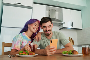 Hipster couple having a breakfast with croissant and salad, taking a selfie. Young woman with colorful hair and bearded man at the kitchen watching memes on the phone. Close up, copy space, background