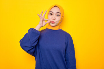Serious young Asian Muslim woman dressed in casual sweater making zipping mouth gesture, keep secret safe isolated over yellow background