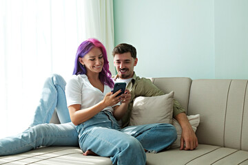 Hipster couple sitting on the couch laughing at memes on the phone at home. Young woman with bright...