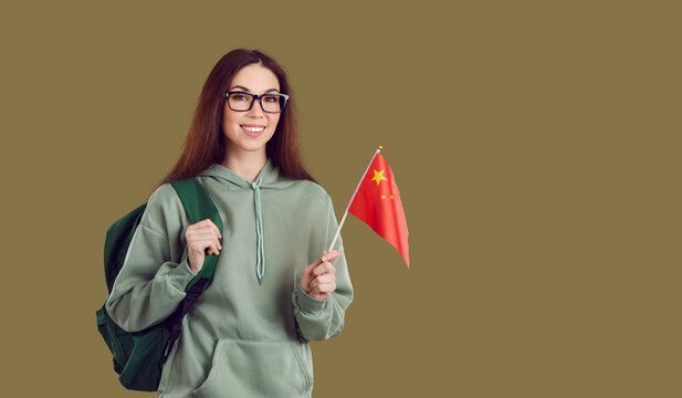 Portrait Of Smiling Millennial Girl In Glasses Isolated On Khaki Green Studio Background Hold Chinese Flag. Happy Student Study Abroad In China Carry Backpack. International Education Concept.