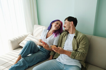 Hipster couple sitting on the couch exchanging memes on their phones at home. Young woman with bright colorful hair and bearded man laughing together. Close up, copy space, background - obrazy, fototapety, plakaty
