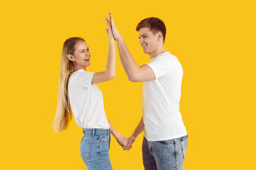 Happy funny couple in love on orange background gives each other high five celebrating success. Smiling young man and woman in jeans and white t-shirts holding hands and touching their palms.