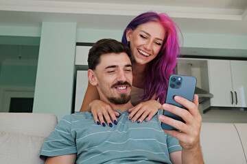 Hipster couple sitting on the couch taking selfies on the phone at home. Young woman with bright colorful hair and bearded man laughing at memes. Close up, copy space, background