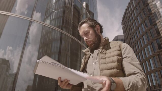 Low angle medium shot of young bearded man in quilted vest looking through business documents while standing near under modern glass office buildings in city downtown on sunny day