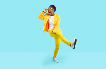 Cheerful young woman having fun, dancing and laughing crazy isolated on light blue background. African American young woman in yellow suit and sunglasses is walking with funny expression on her face.