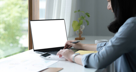 Mockup image of a woman using laptop with blank white screen on wooden table in office