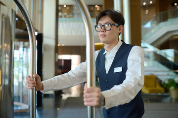 Young man in uniform of bellboy pushing cart with luggage of guests of hotel while moving along...