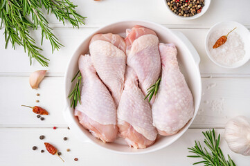 Raw uncooked chicken legs in a white dish on a white wooden background. Meat with ingredients for...