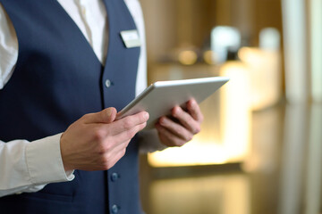 Digital tablet in hands of young male receptionist of luxurious modern hotel standing in lounge by...