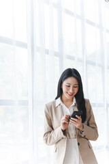 Portrait of elegant successful asian business woman speaking by smartphone and smiling happily while standing against window in office.