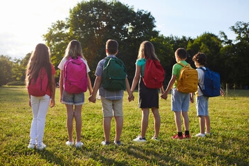 Schoolchildren with backpacks on their shoulders, holding hands, stand in row in park. Back view of...
