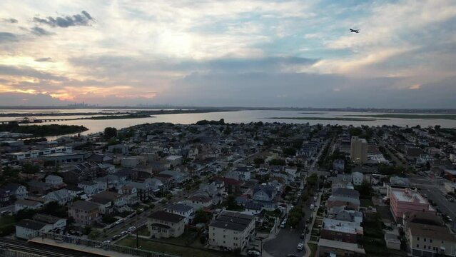 An aerial view over Arverne, NY during a cloudy but beautiful sunset. The camera truck right and pan left above apartment buildings following a plane and the sunset in the distance on the horizon.
