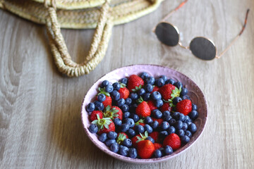 Bowl of strawberries and blueberries, sunglasses and wicker bag on the wooden table. Selective focus. 