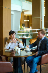 Two young interracial businessmen in formalwear having coffee in cafe or lounge of modern hotel and discussing working points