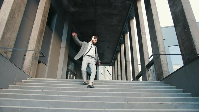 Young Caucasian employee in suit listening to music in headphones, holding smartphone, dancing while walking down stairs backdrop office building. Concept of happiness, good mood, success