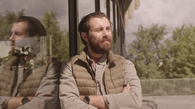 Waist up portrait of young Caucasian bearded man in shirt and quilted vest looking around and at camera, standing with hands folded in city downtown leaning on glass office building