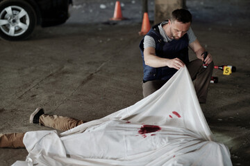 Young policeman or criminological expert in bulletproof vest inspecting corpse of killed person...