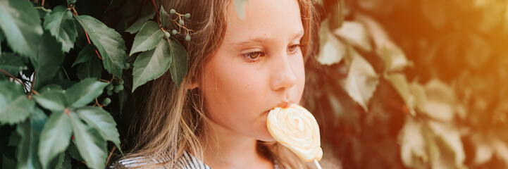 candid thoughtful little kid girl of eight years old holding lollipop and lick eating fruit sugar sweet candy on the background of hedge wall of green plants in summer vacation outdoor. banner. flare - obrazy, fototapety, plakaty