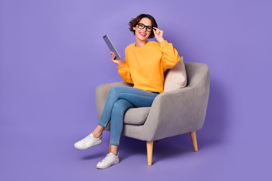 Portrait of attractive cheerful girl sitting using device reading e book touching specs isolated over violet lilac color background