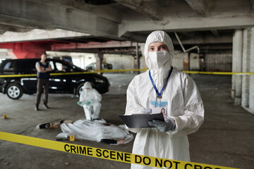 Young female crime scene investigator in protective coveralls making notes in document while...