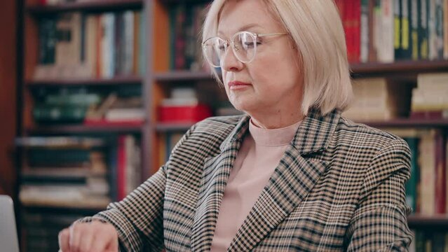 Senior female professor in eyeglasses working on laptop at college library