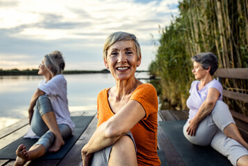 Group of senior woman doing yoga exercises by the lake. - obrazy, fototapety, plakaty