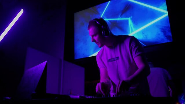 DJ work in club in neon lights indoors. Caucasian male person using controller console to mix music and change volume sound. Purple light in nightclub room. Jockey on disco party. Nightlife in city