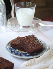 An angle view shot of plate of fluffy and crunchy pieces of dark chocolate brownie with a glass of milk in background. It is a square or rectangular chocolate baked