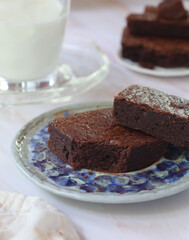 A plate of fluffy and crunchy pieces of dark chocolate brownie with a glass of milk in background. It is a square or rectangular chocolate baked confection
